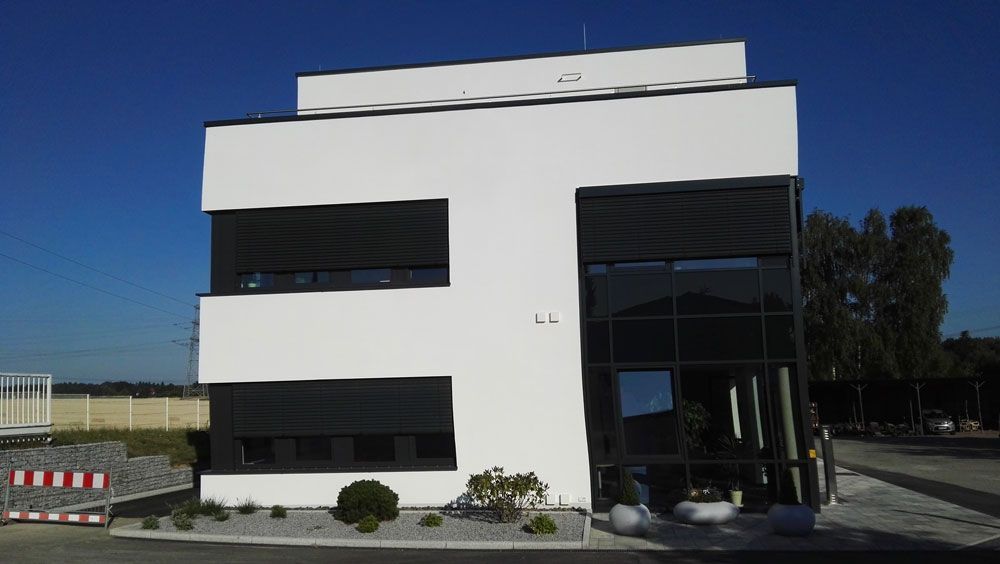 Weißes, modernes Gebäude mit dunklen Fensterverkleidungen vor blauem Himmel.