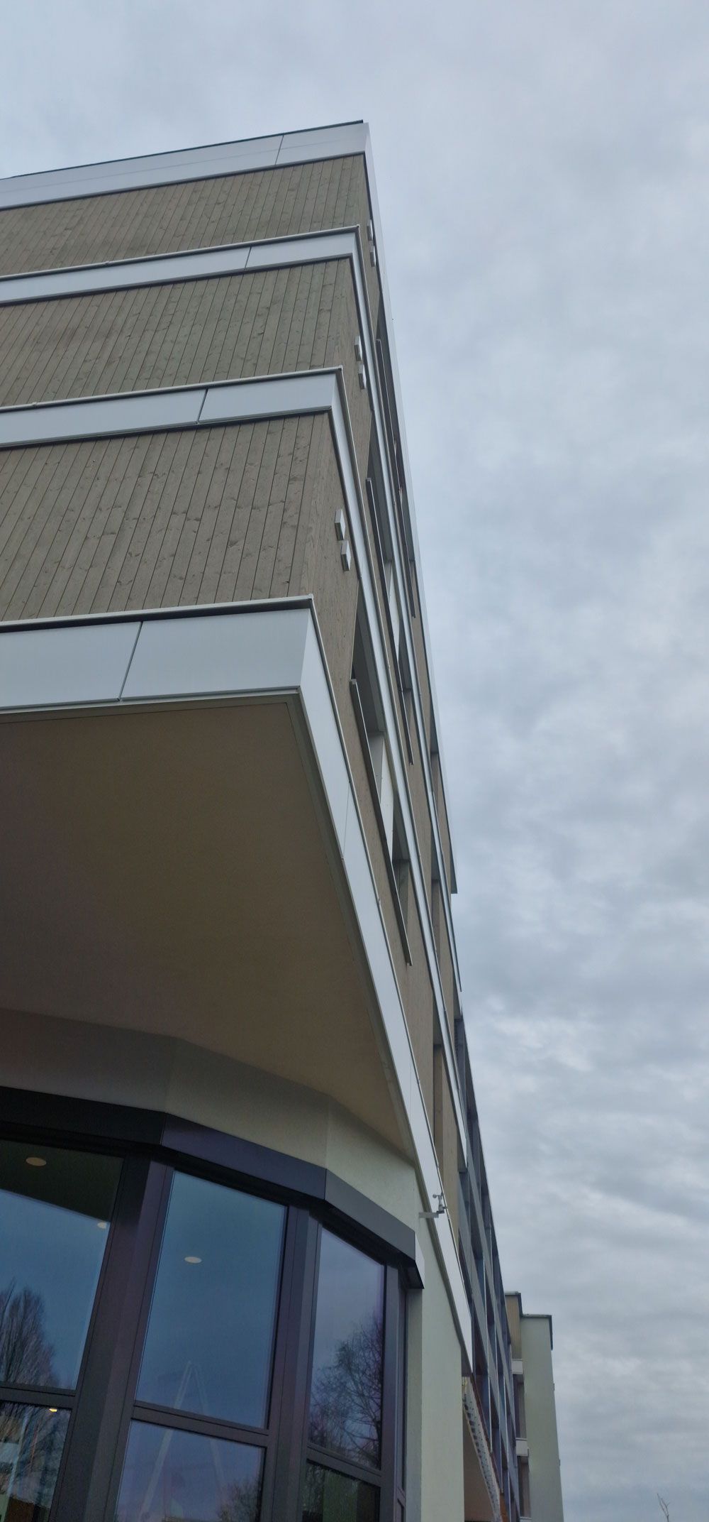 Ansicht von unten auf ein modernes Gebäude mit abgerundeten Ecken, beiger Fassade und Glasfenstern. Bewölkter Himmel.