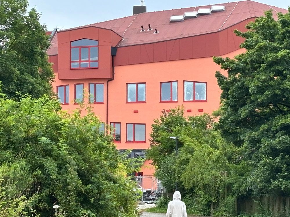 Ein orangefarbenes Gebäude mit rotem Dach, von hinter grünen Bäumen aus gesehen; eine Person in Weiß geht auf das Gebäude zu.