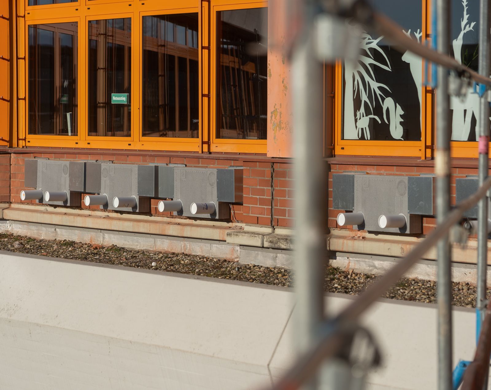 Außenansicht eines Gebäudes mit orangefarbenen Holzfenstern und grauen Steinblöcken. Im Vordergrund ist ein Gerüst zu sehen.