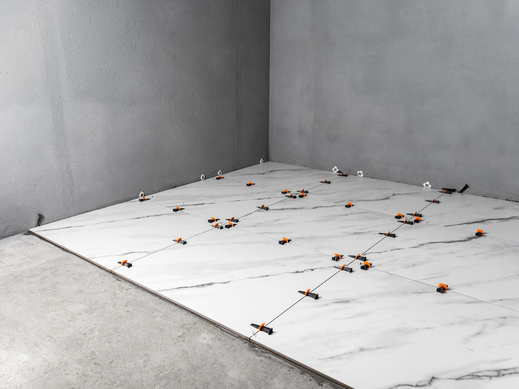 Pose de grands carreaux blancs à motif marbre sur un sol en béton, avec des cales de nivellement.