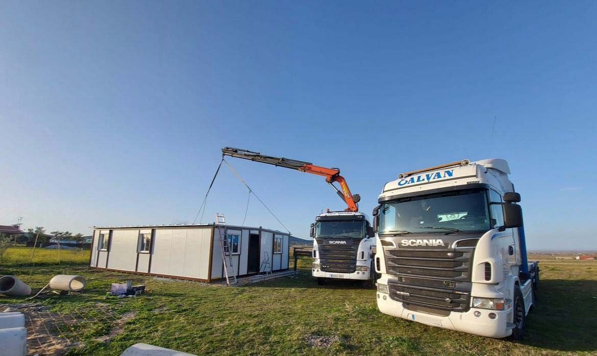 Dos semirremolques están estacionados en un campo con una grúa levantando un edificio.
