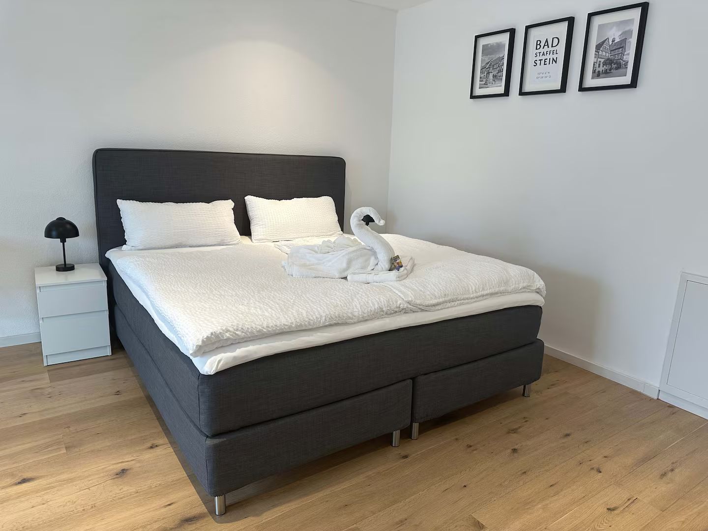 Ordentliches Schlafzimmer mit grauem Bett, weißer Bettwäsche, Nachttisch und gerahmten Bildern an weißen Wänden.