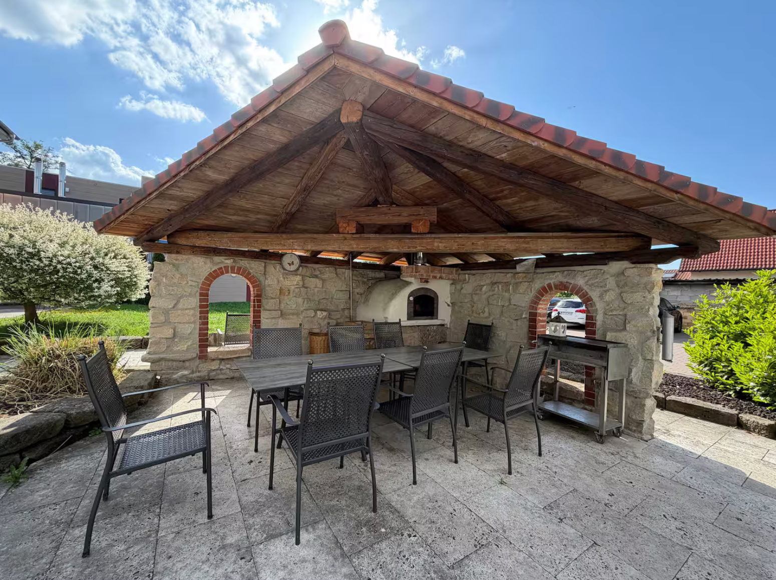 Steinpavillon im Außenbereich mit Holzdach, Terrassensitzgelegenheiten und eingebautem Grill in einem Garten.