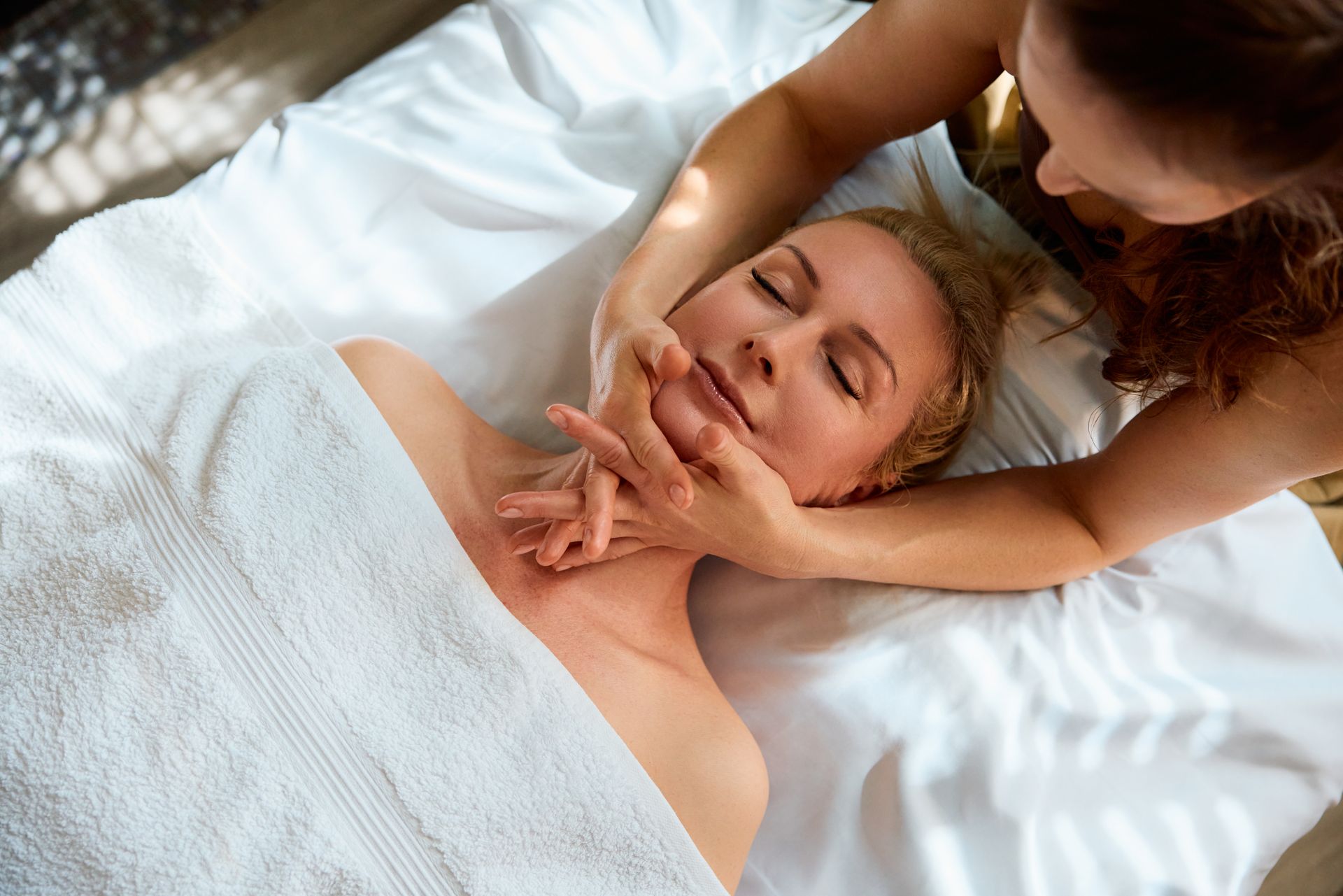 Femme recevant un massage du visage sur une table de massage. Mains sur le visage et le cou ; serviette blanche.