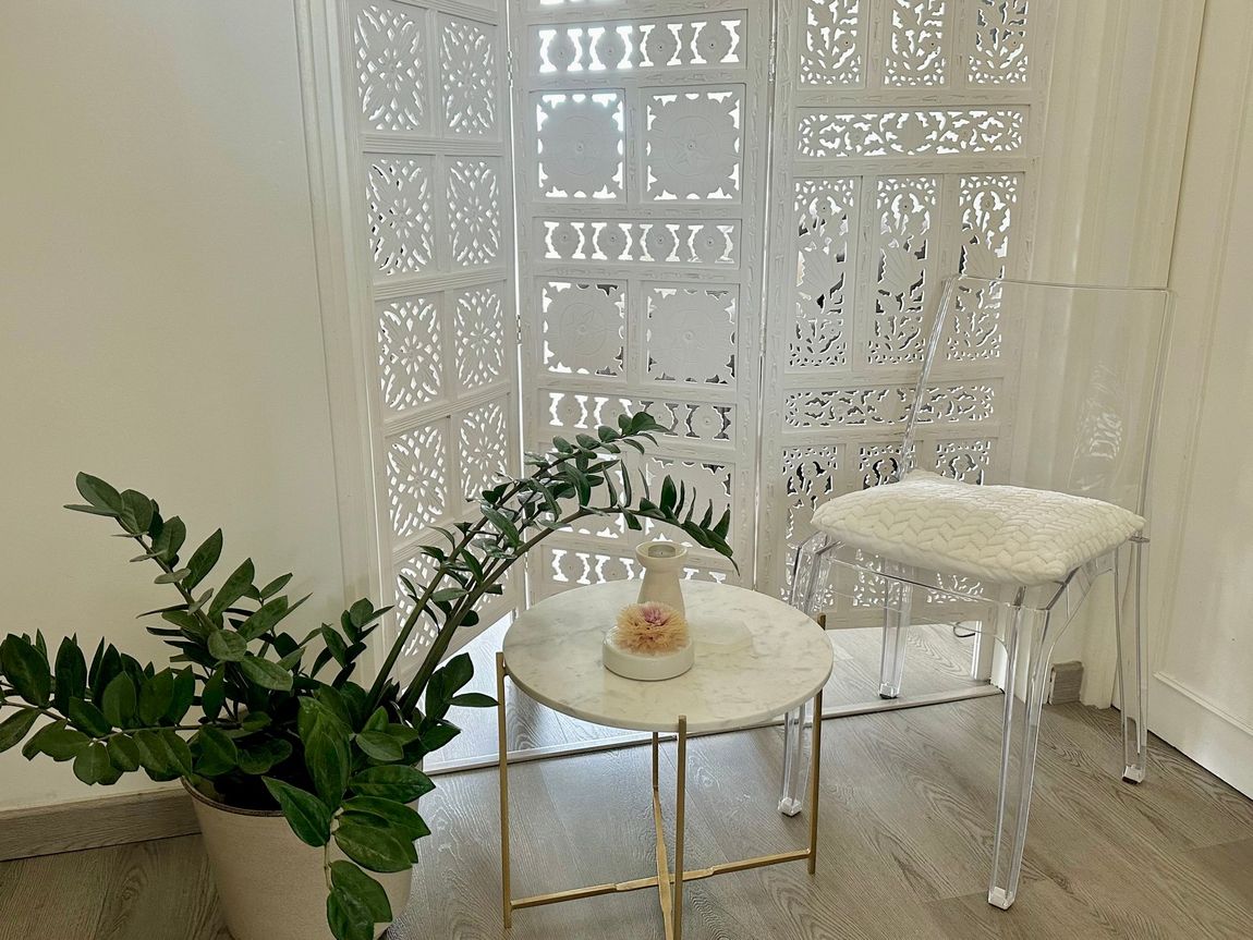Pièce blanche avec un paravent décoratif, une chaise transparente, une petite table avec un vase et une plante en pot.