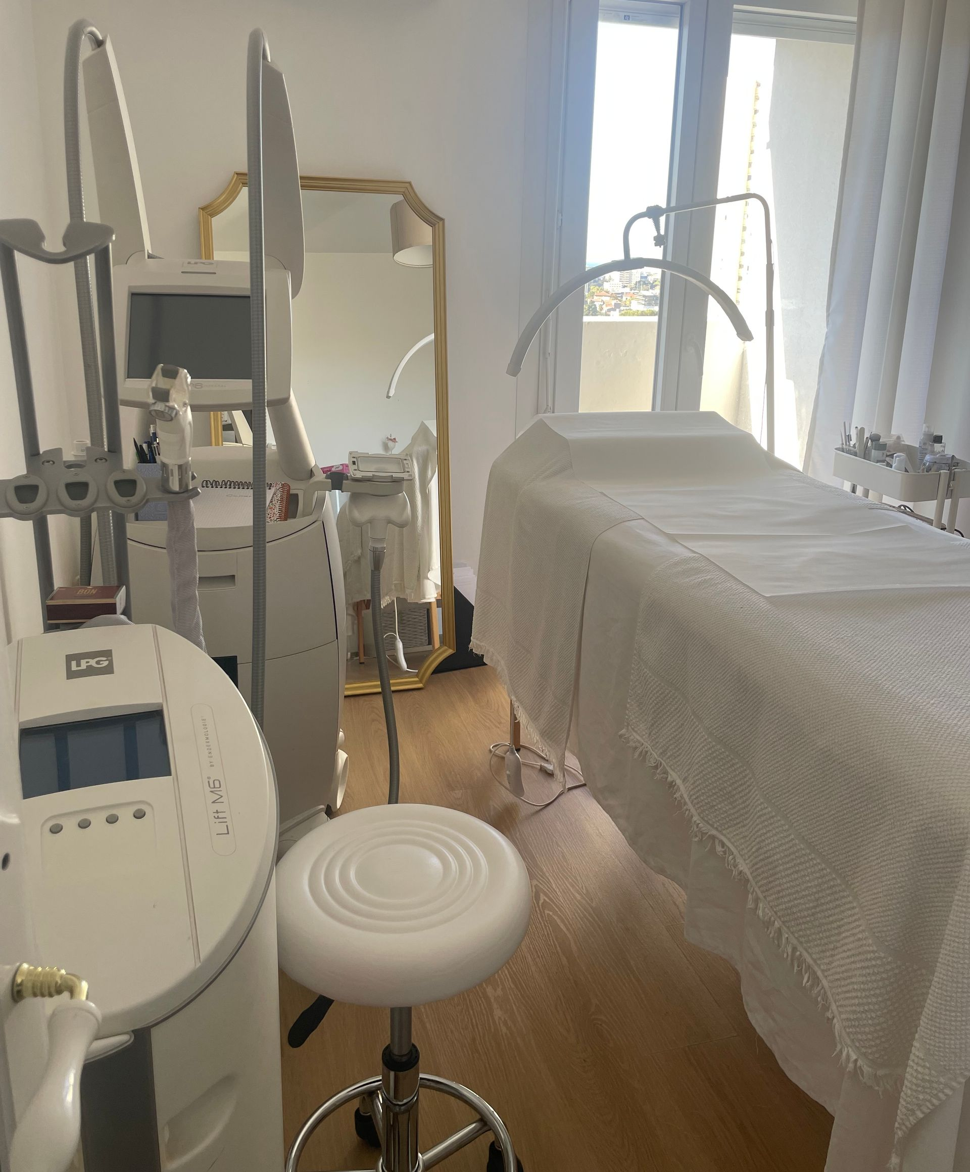 Salle de soins d'esthétique. Un lit, du matériel médical, un tabouret et un grand miroir sont présents dans la pièce.