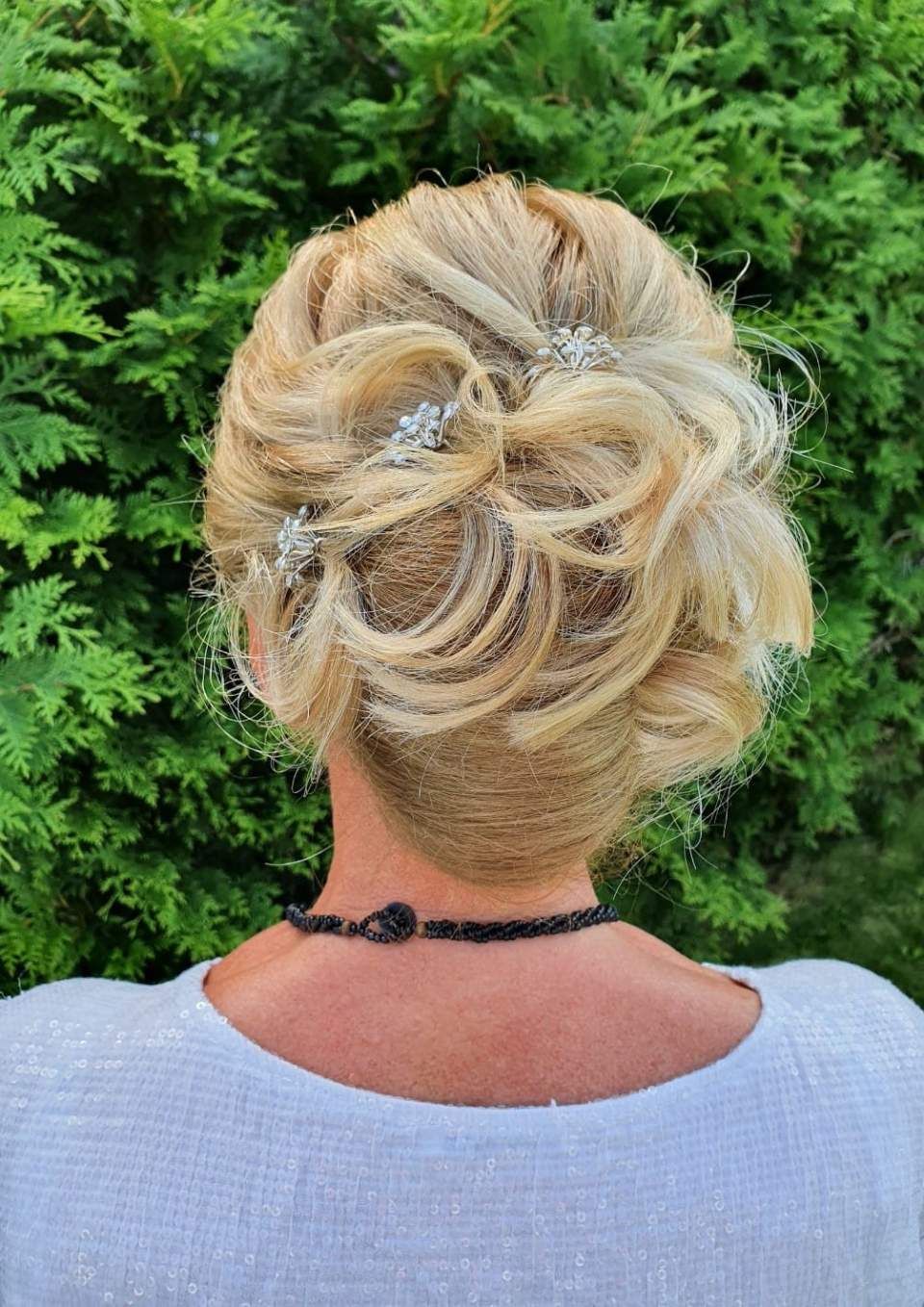 Blonde Hochsteckfrisur mit Locken und Schmuckakzenten; vor grünem Hintergrund.