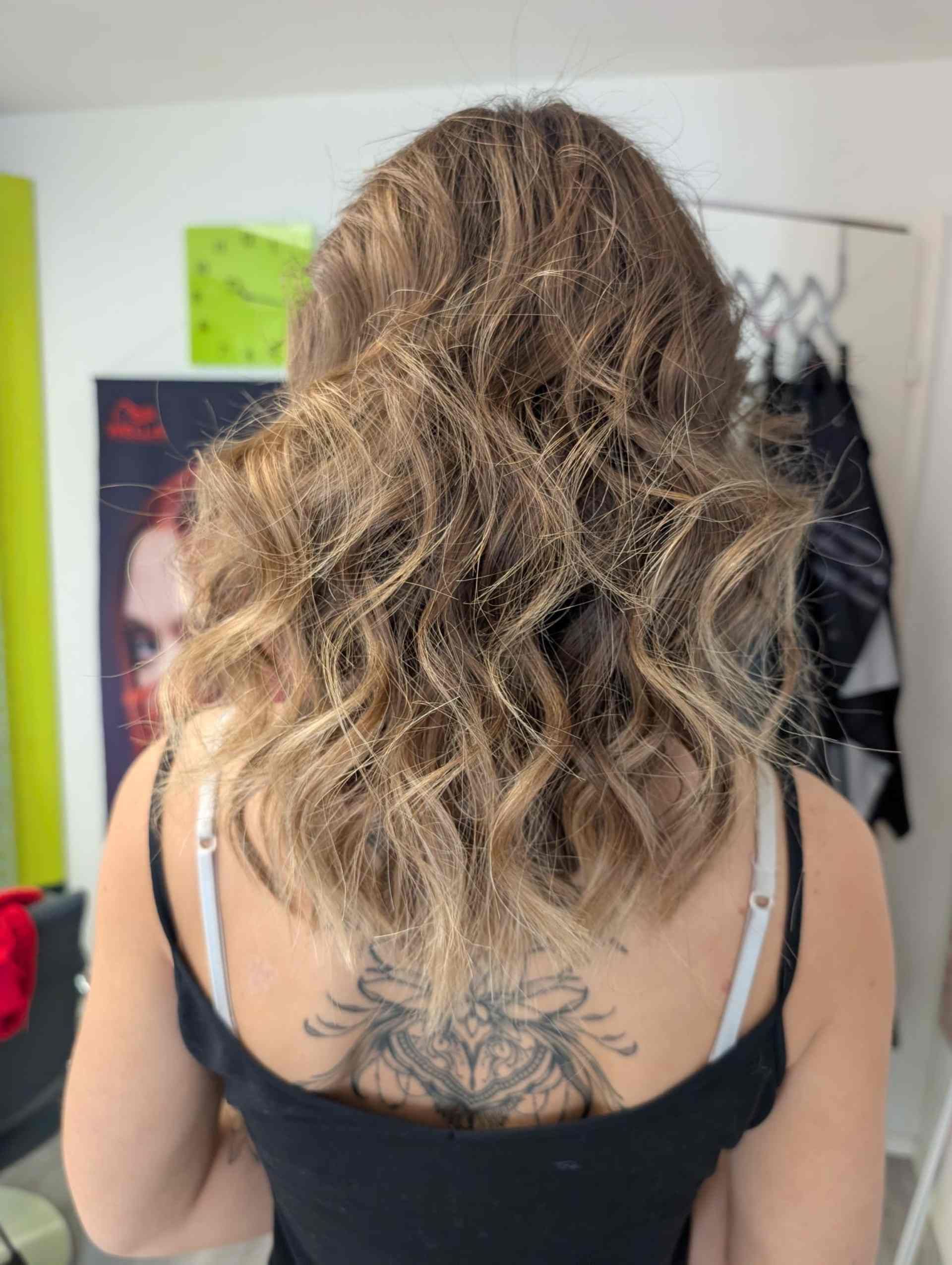 Frau mit lockigem, gestuftem braun-blondem Haar, mit dem Rücken zur Kamera, trägt ein schwarzes Tanktop.