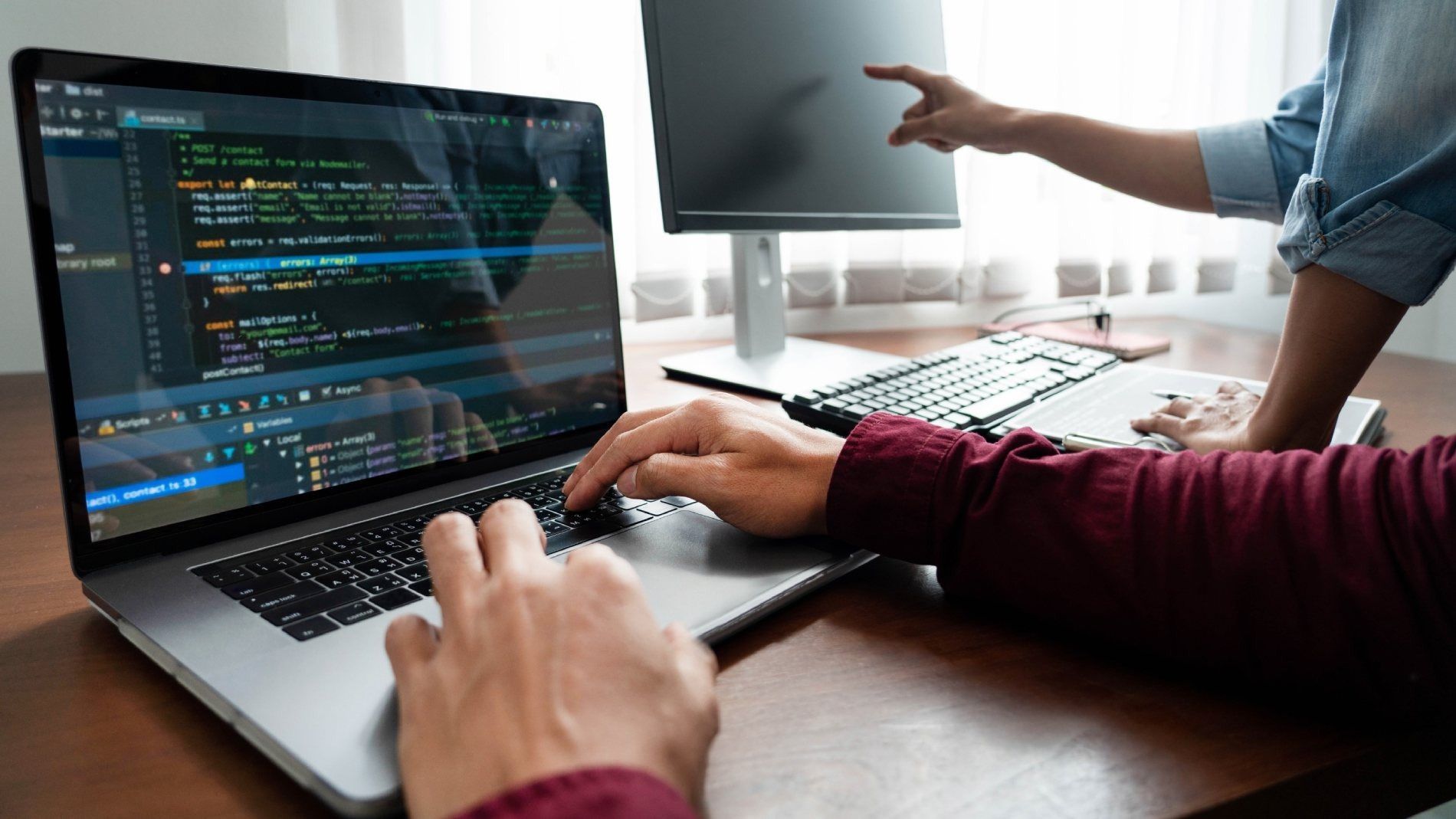 Una persona está escribiendo en una computadora portátil mientras otra persona señala una pantalla de computadora.