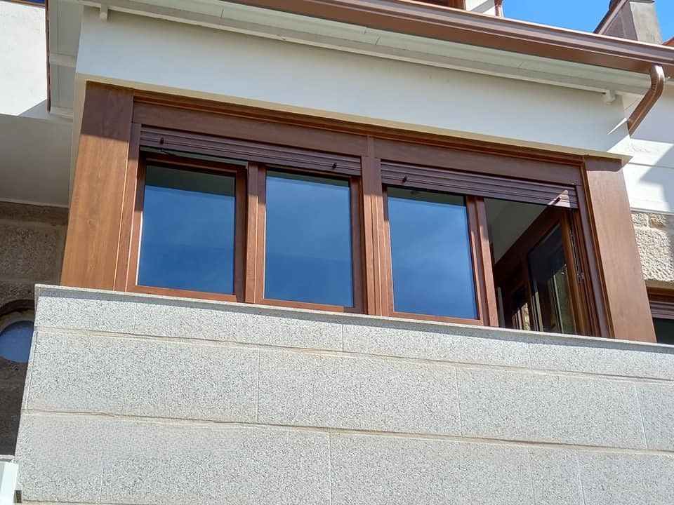 Tres ventanas de marco oscuro y contraventanas marrones en un edificio de color claro, sobre un muro de piedra.