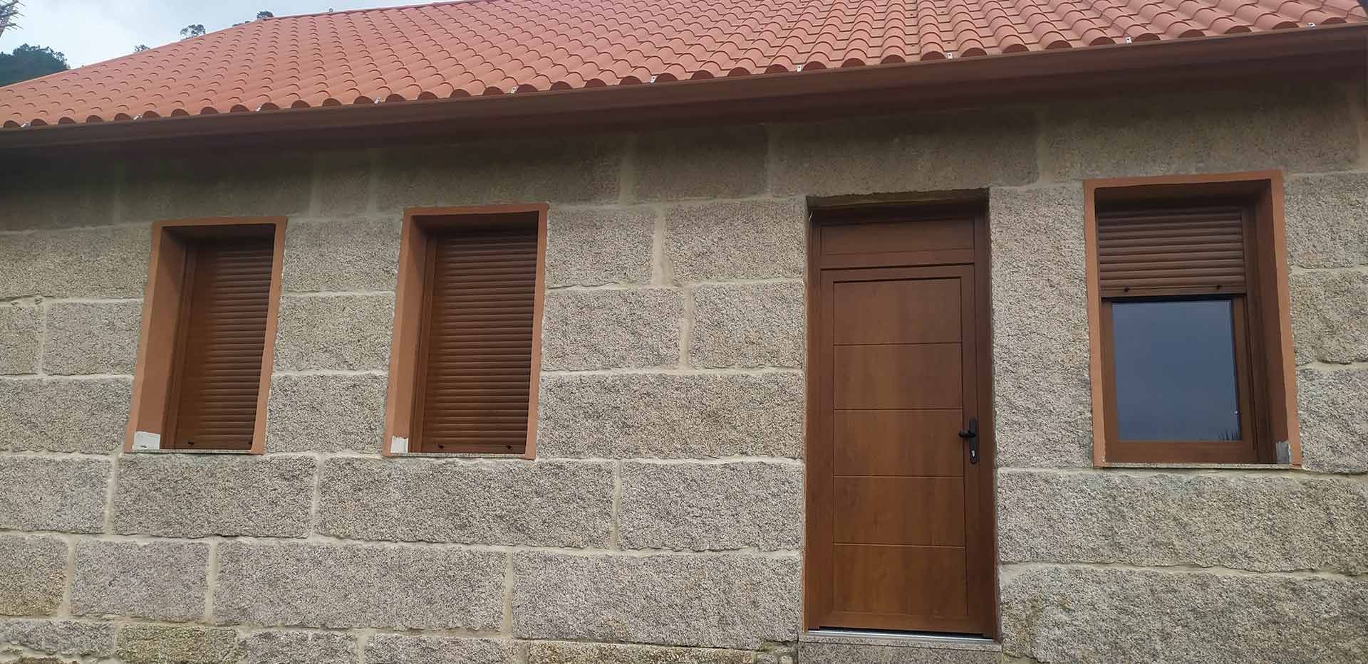 Edificio de piedra con puerta marrón, ventanas con contraventanas y techo de tejas rojas.