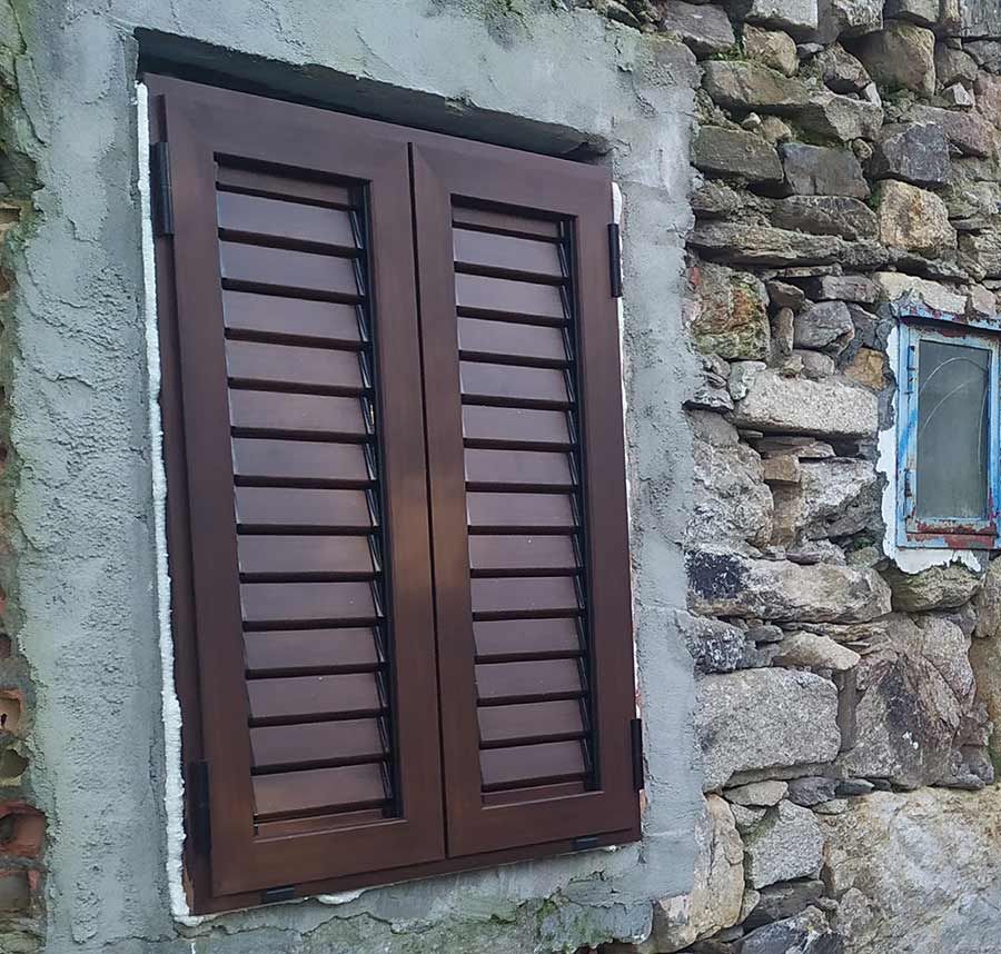 Ventana con contraventanas marrones en una pared de piedra, al lado de una ventana más pequeña con marco azul.