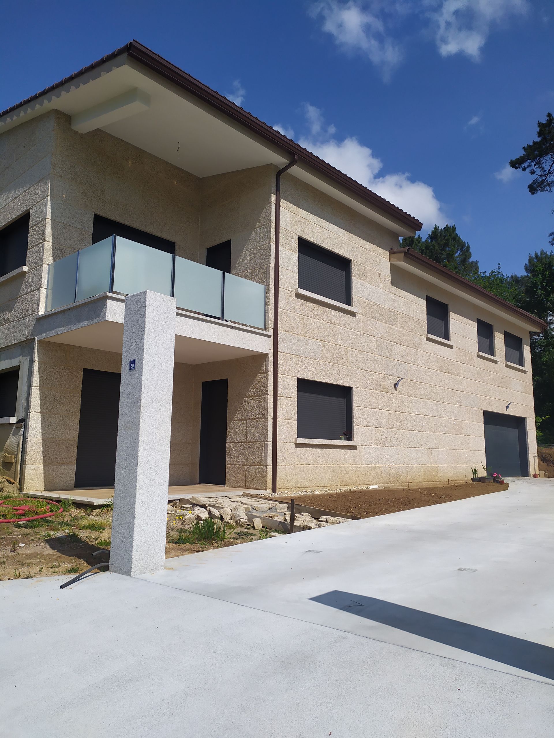 Casa de piedra de dos pisos con entrada de concreto y balcón, bajo un cielo azul.