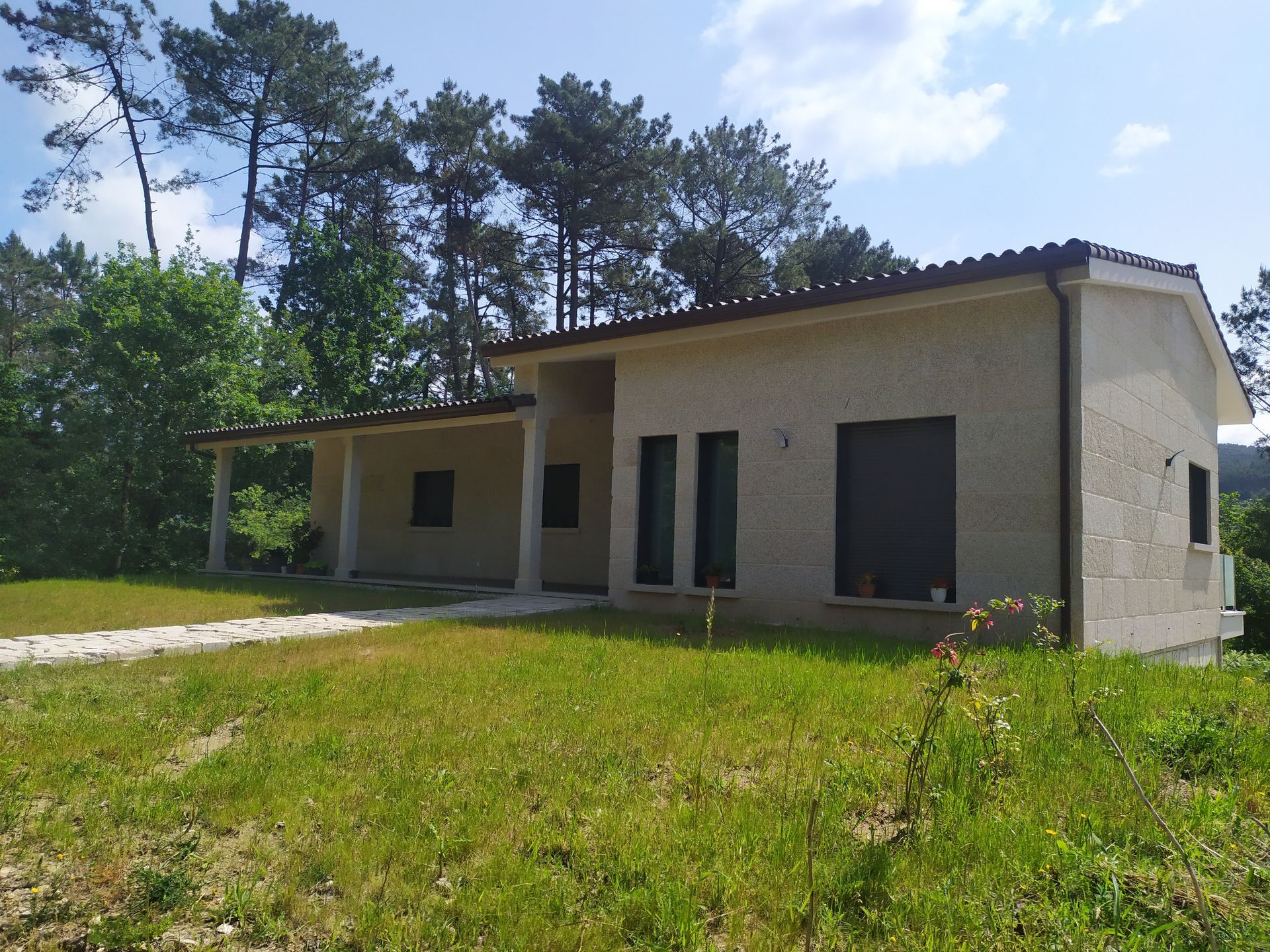 Una casa beige de una sola planta con un porche cubierto en una zona verde, árboles al fondo, bajo un cielo azul.