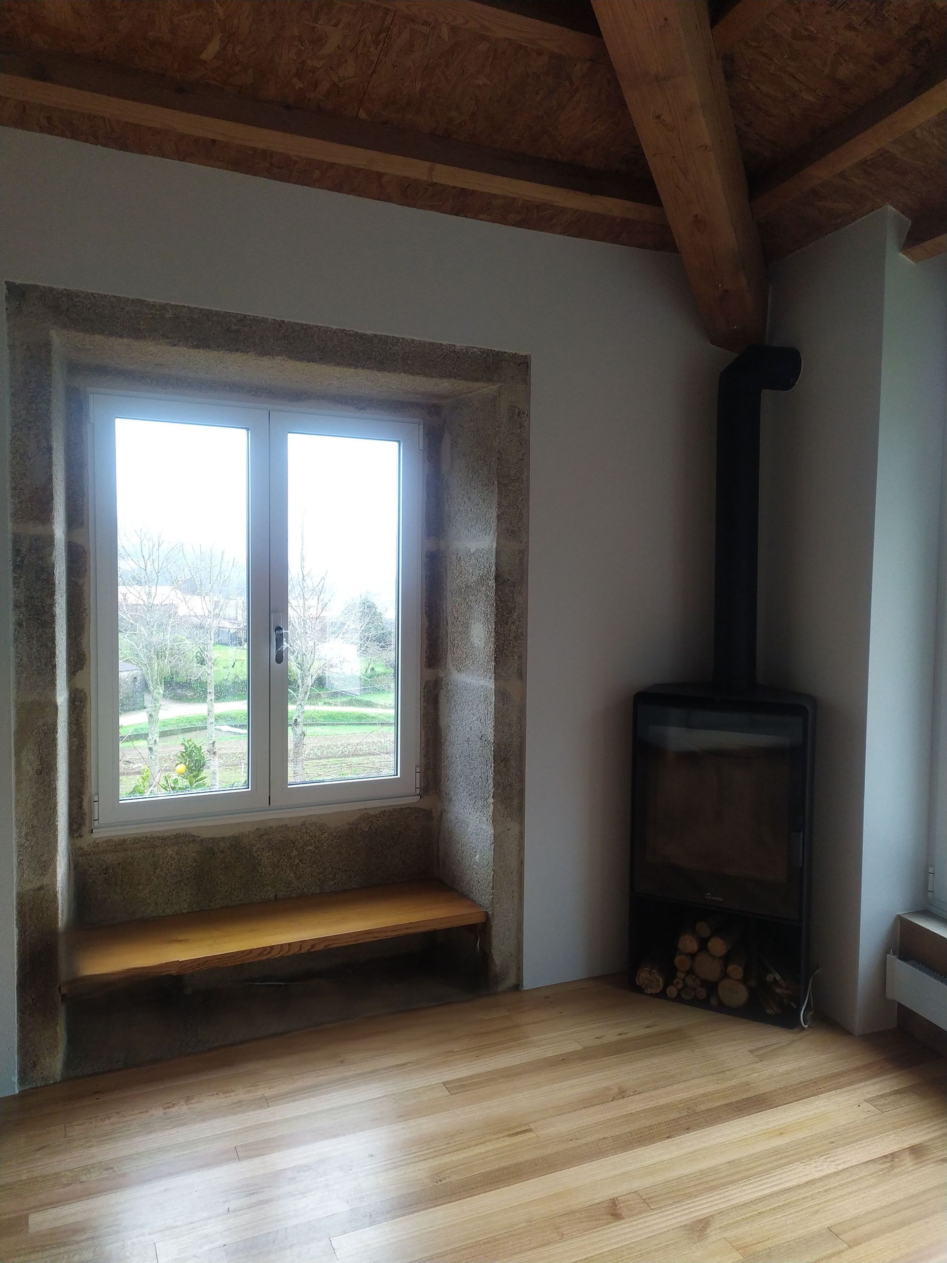 Habitación con ventana y estufa de leña. Marco de piedra, suelo de madera clara y vigas de madera a la vista.