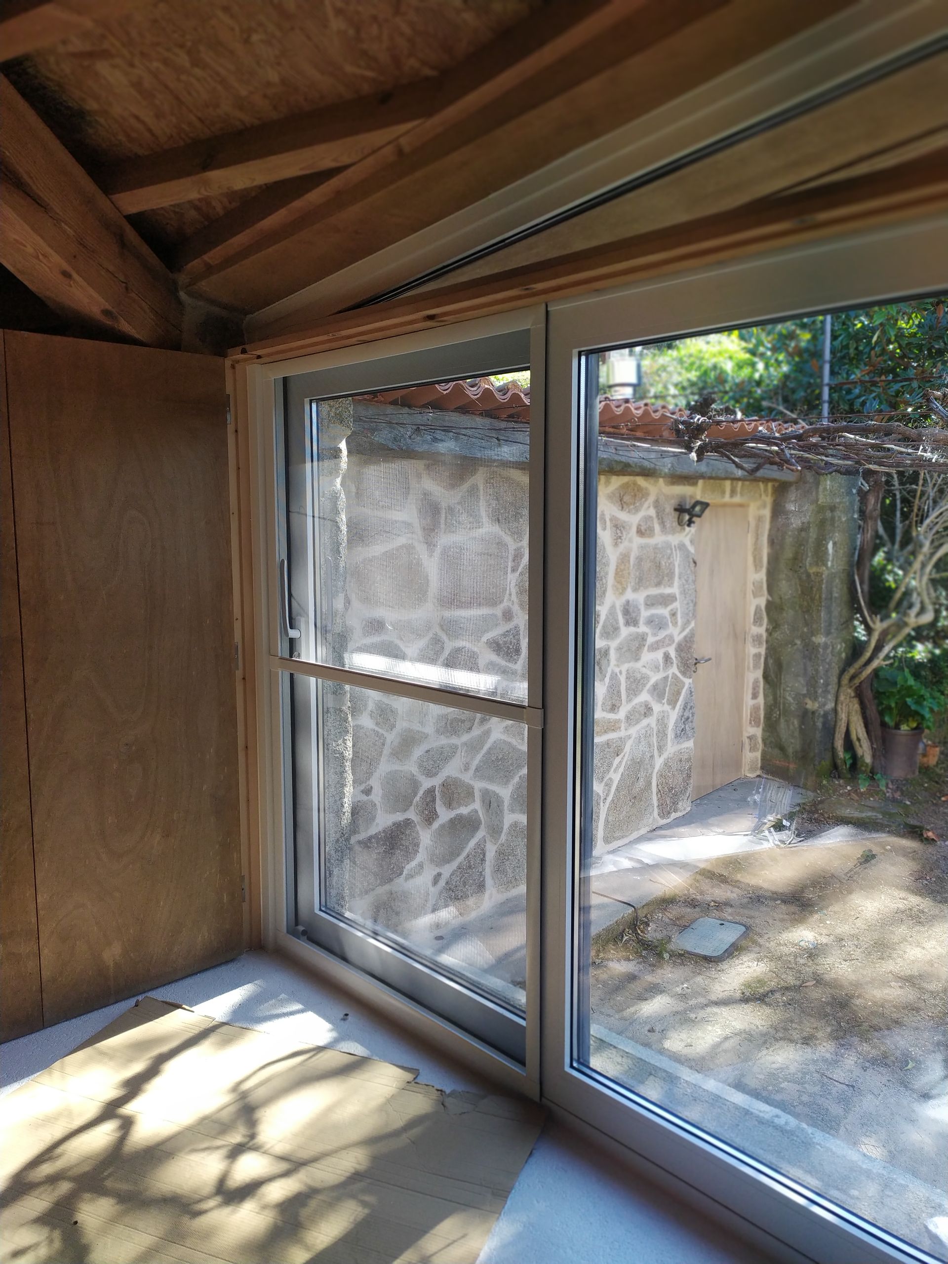 Puerta corrediza de vidrio en una habitación con un muro de piedra visible al exterior. Vigas de madera y pared sin terminar.