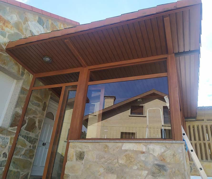 Porche de madera marrón con ventanas, luz y un muro de piedra. El cristal reflectante muestra otra casa.
