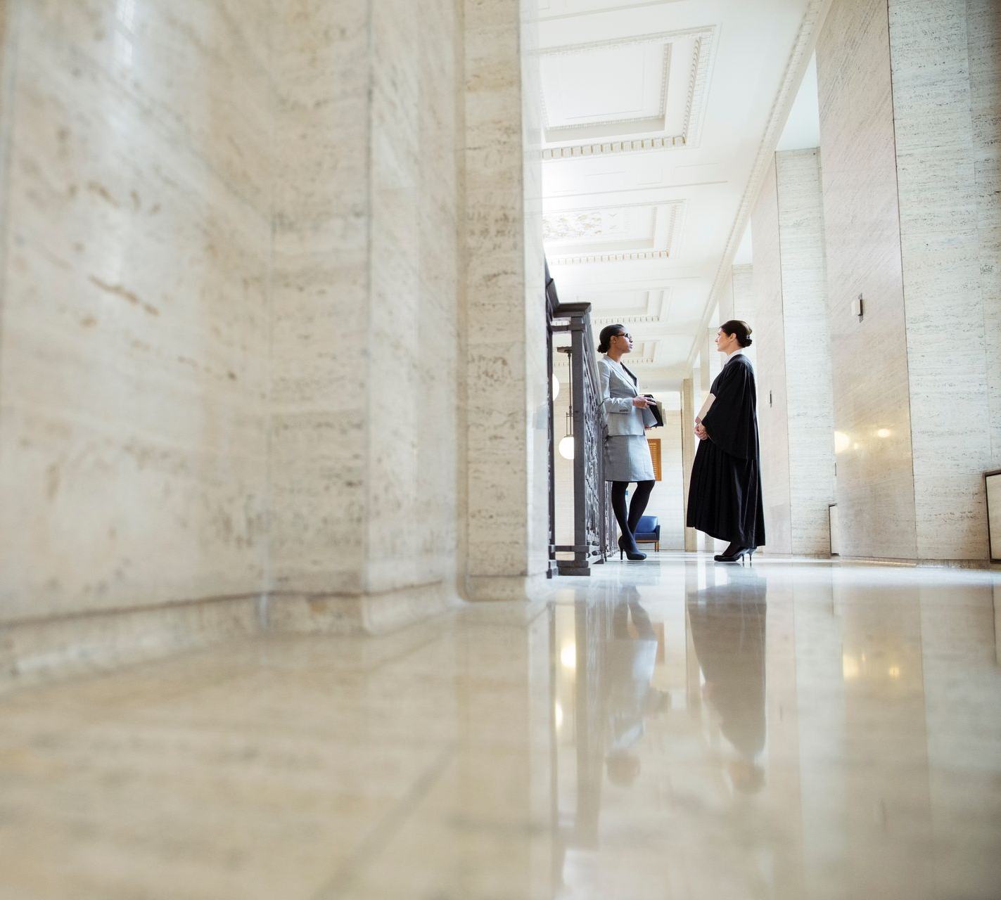 Deux personnes dans un couloir en marbre, levant les yeux. L'une porte un manteau, l'autre une robe sombre.