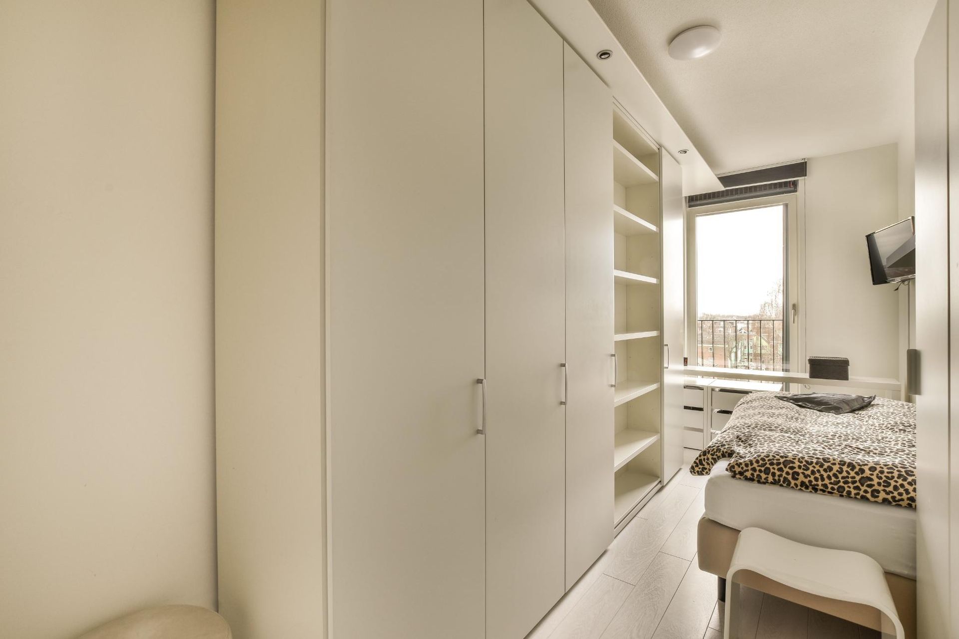 Habitación luminosa y minimalista con armarios empotrados blancos, una cama y vistas al balcón a través de la ventana.