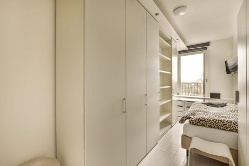 Habitación luminosa y minimalista con armarios empotrados blancos, una cama y vistas al balcón a través de la ventana.