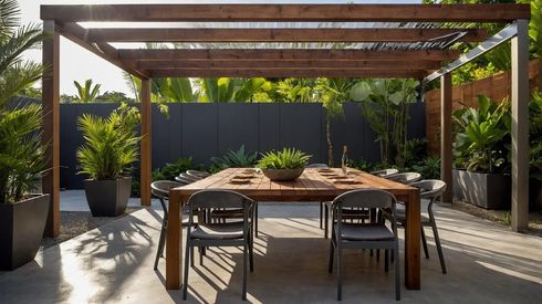 Comedor exterior en el patio con mesa de madera, sillas, pérgola y plantas en macetas, bañado por la luz del sol.