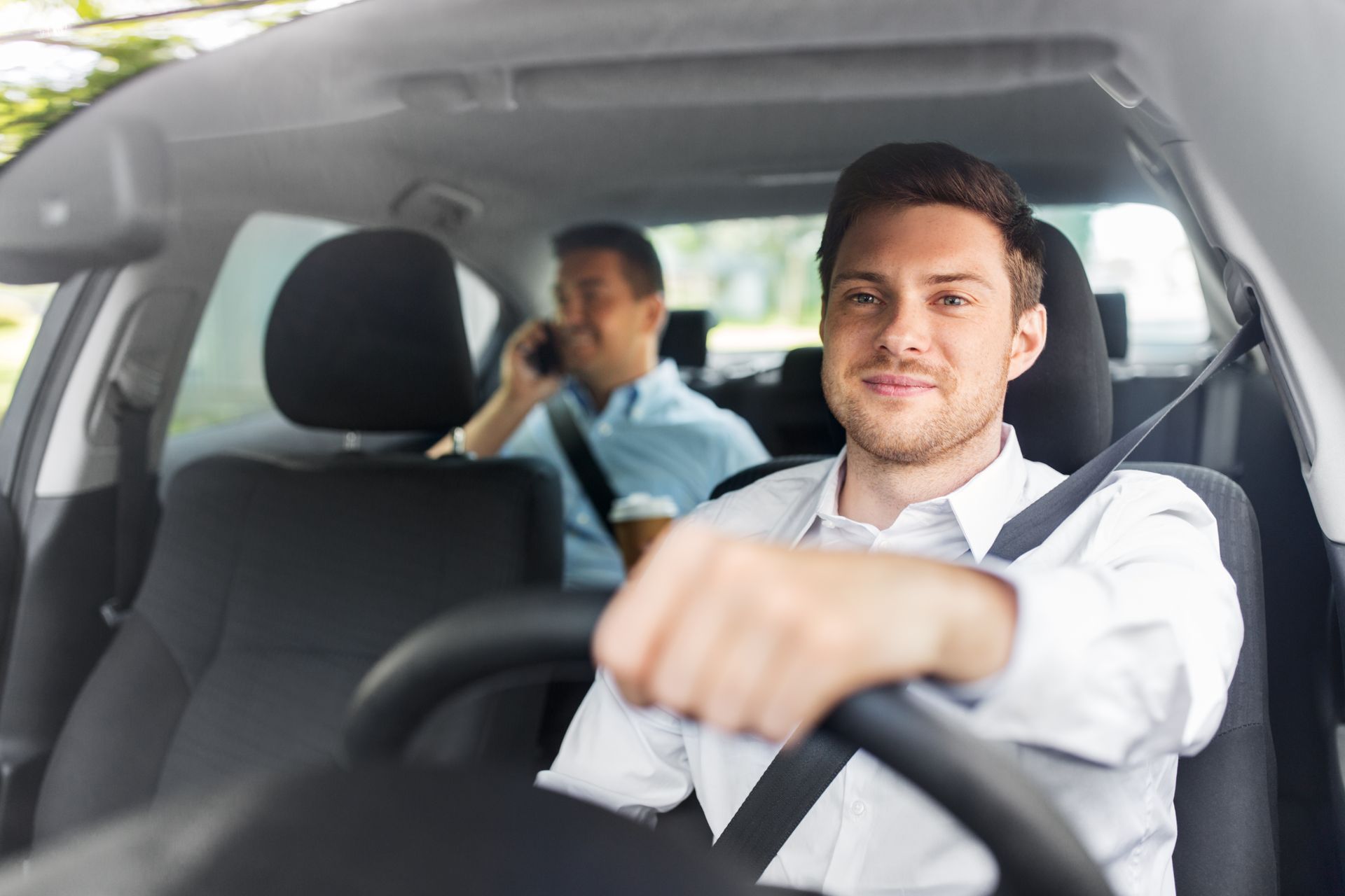 Un homme conduit une voiture, souriant, avec un passager à l'arrière au téléphone.