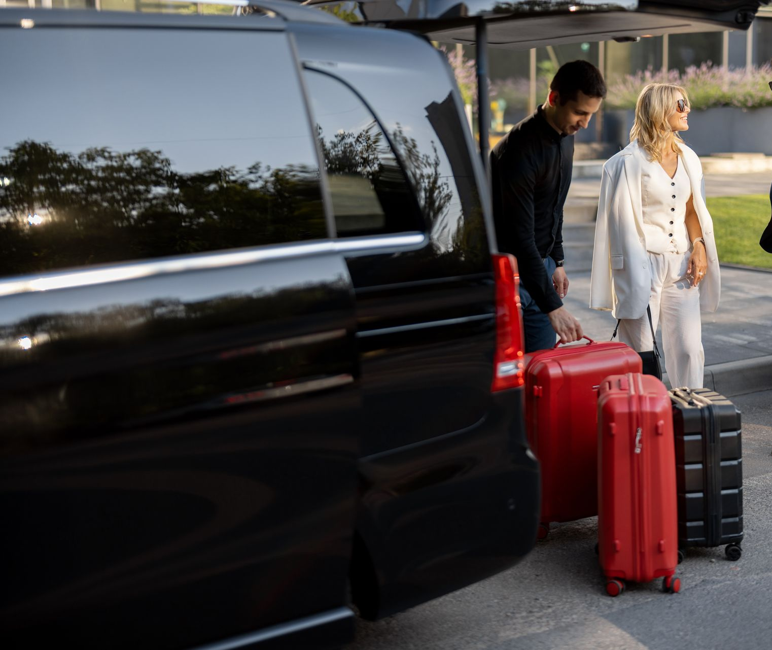 Un homme charge des bagages dans une camionnette noire ; une femme observe à proximité. Valises rouges et noires. En extérieur.