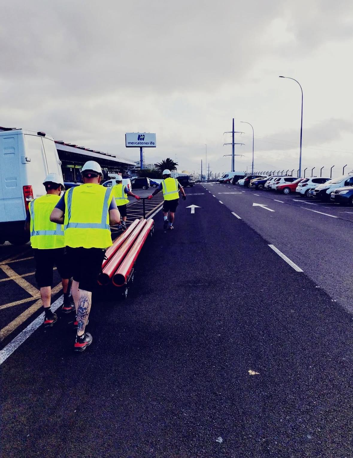 Trabajadores con chalecos reflectantes transportan tuberías largas a lo largo de una carretera mojada bajo un cielo gris.