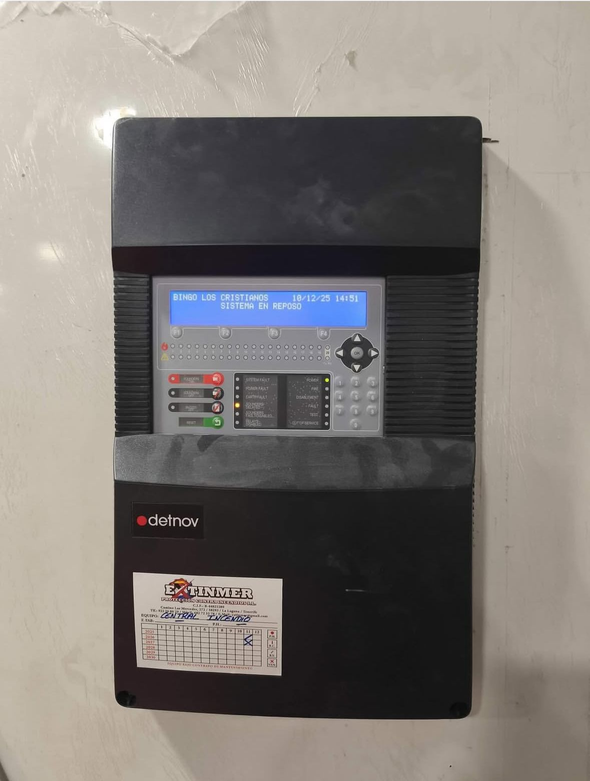 Panel de control eléctrico negro montado en la pared, con pantalla y botones azules, sobre una pared blanca.
