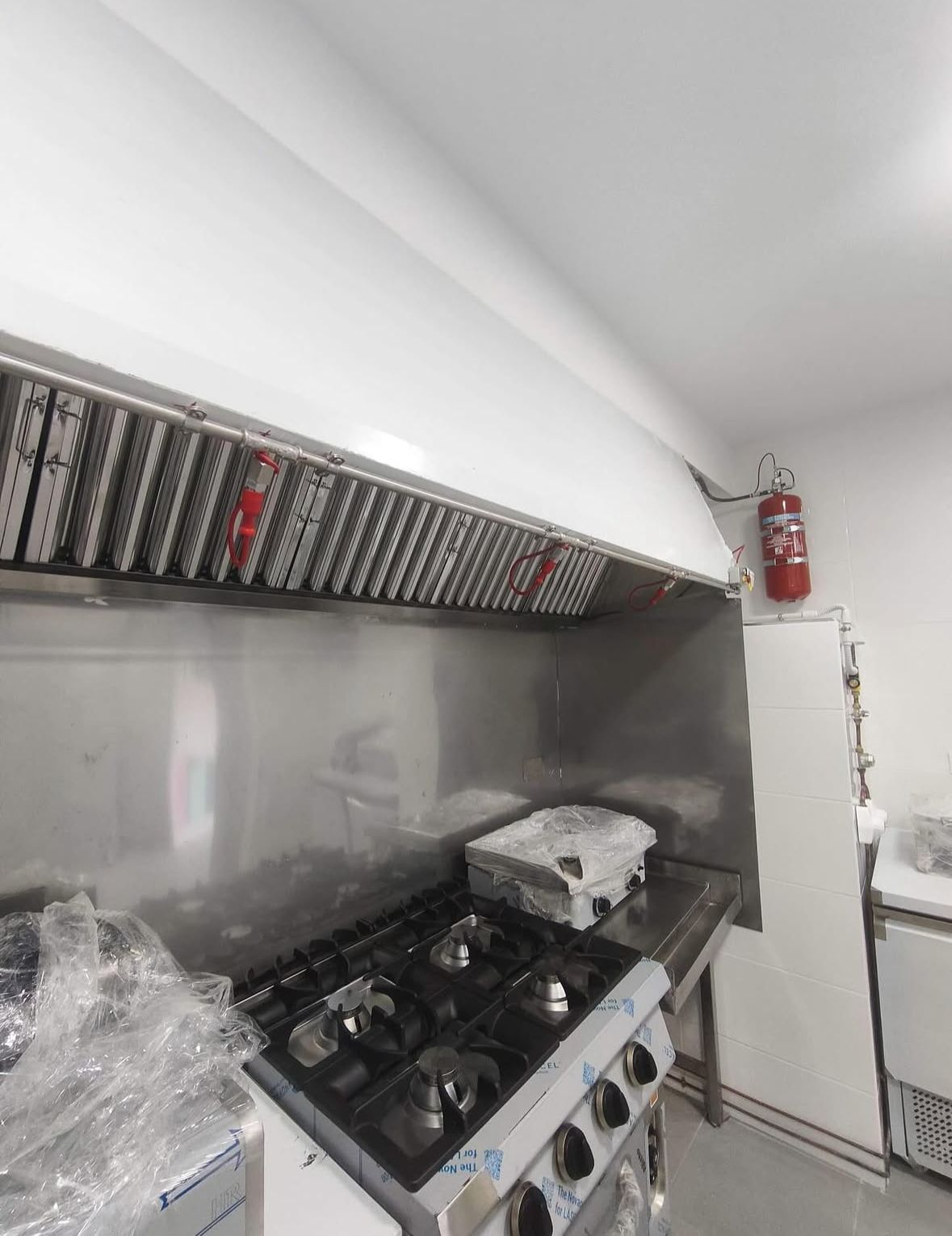 Cocina comercial con estufa de acero inoxidable, campana extractora y extintor rojo en la pared.