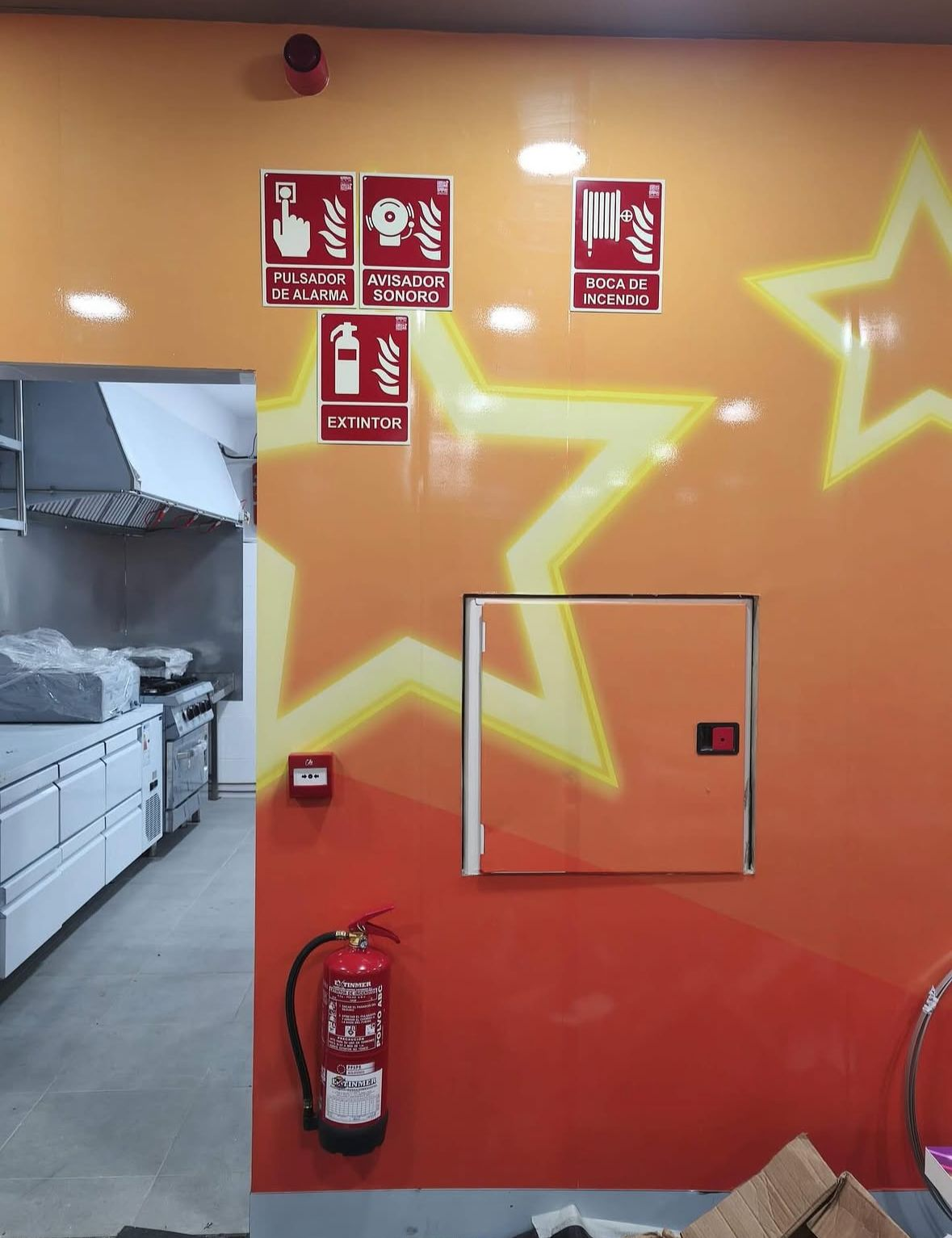 Pared naranja con señales de emergencia, extintor de incendios, panel de acceso y puerta de entrada a una zona de cocina.