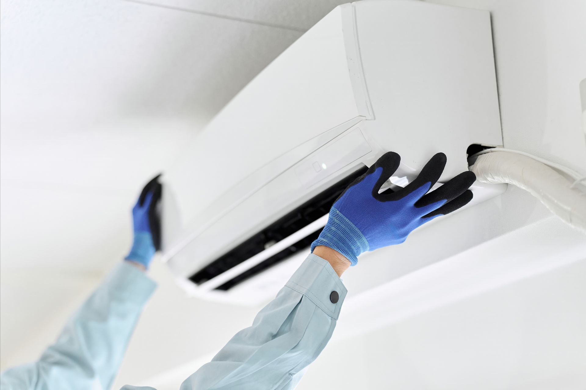 Un technicien portant des gants bleus nettoie un climatiseur mural de plafond.