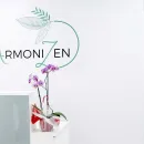 Logotipo con una hoja verde y texto sobre una pared blanca, encima de una orquídea morada en un jarrón blanco.