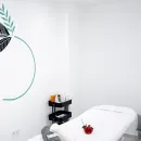 Sala de tratamientos de spa minimalista con camilla de masaje blanca, estante negro y rosa roja sobre la mesa.