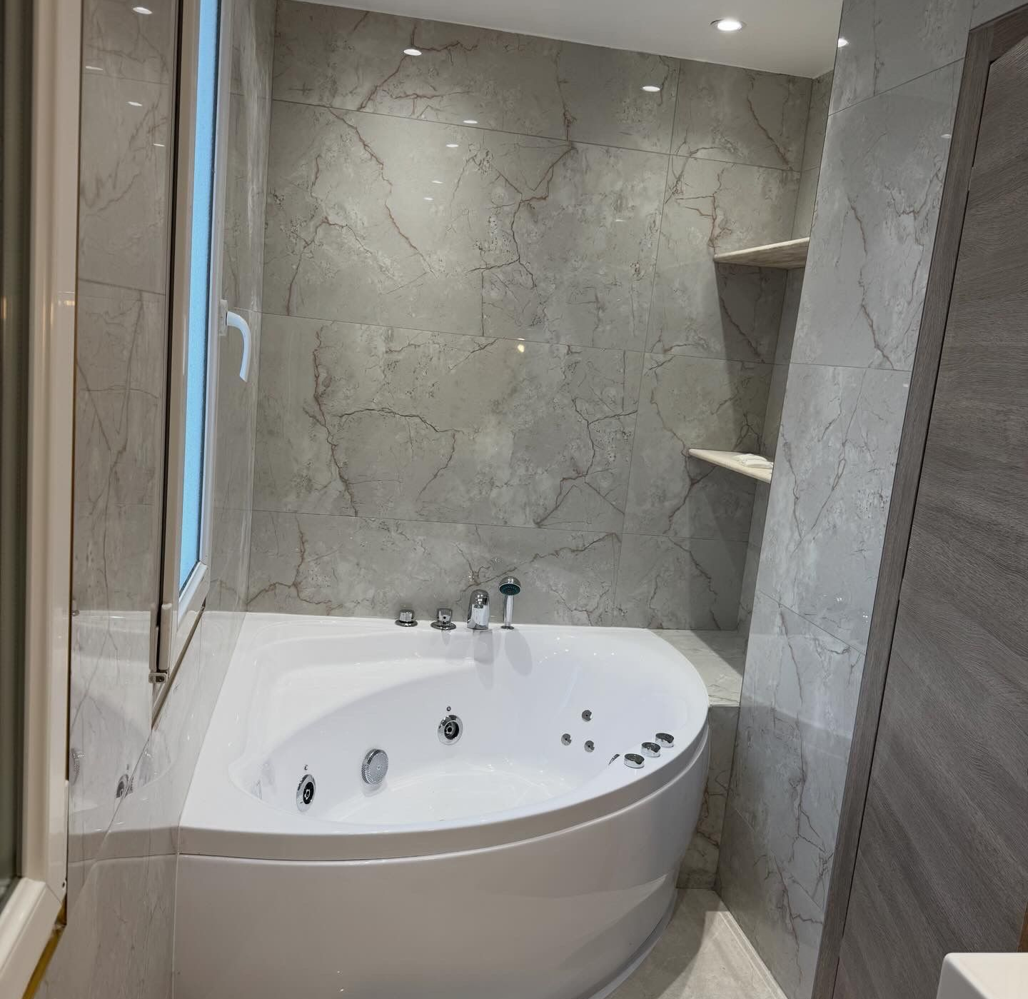 Salle de bains moderne avec baignoire balnéo d’angle, murs en pierre et cabine de douche vitrée, éclairée par des spots encastrés.