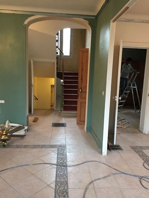 Couloir intérieur en rénovation, avec des murs, un sol carrelé et des ouvriers sur une échelle en arrière-plan.