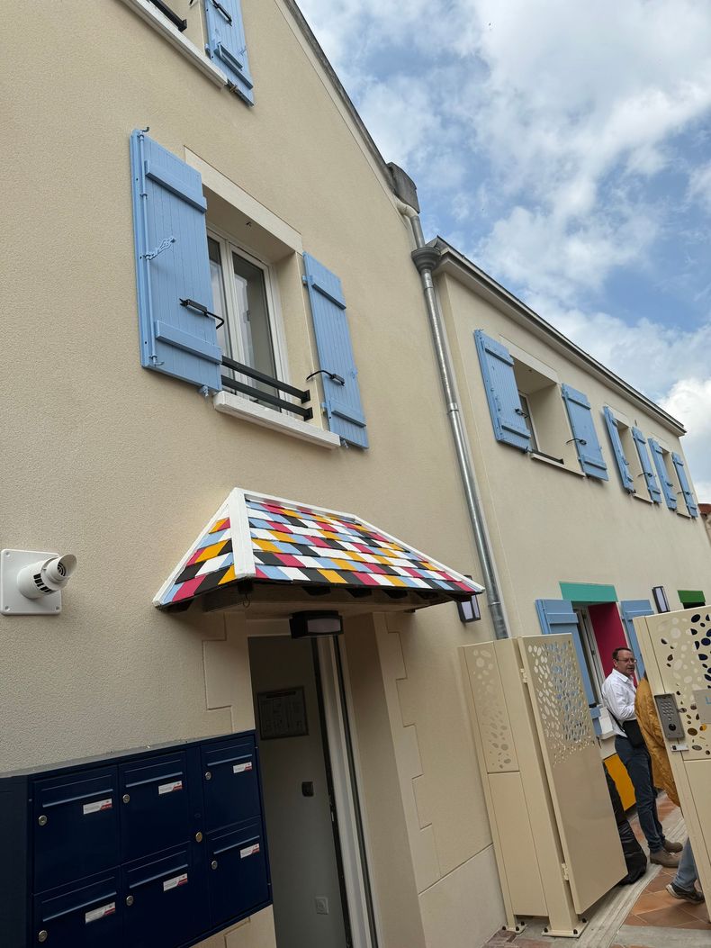 Maison à deux étages, aux volets, avec une marquise au-dessus de la porte d’entrée ; une personne se tient près de l’entrée.