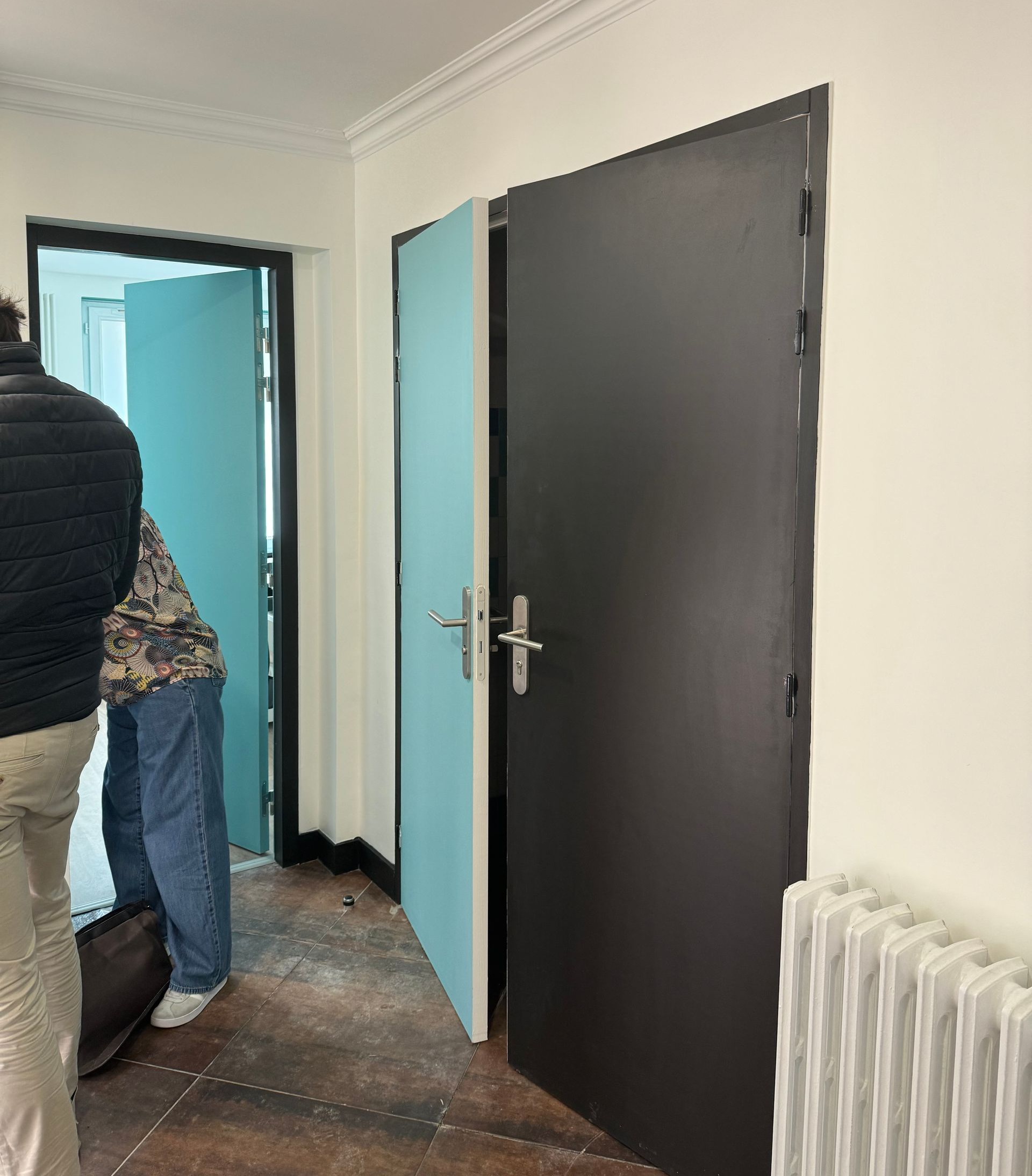 Couloir avec une porte ouverte, une porte fermée, un radiateur et deux personnes à gauche.