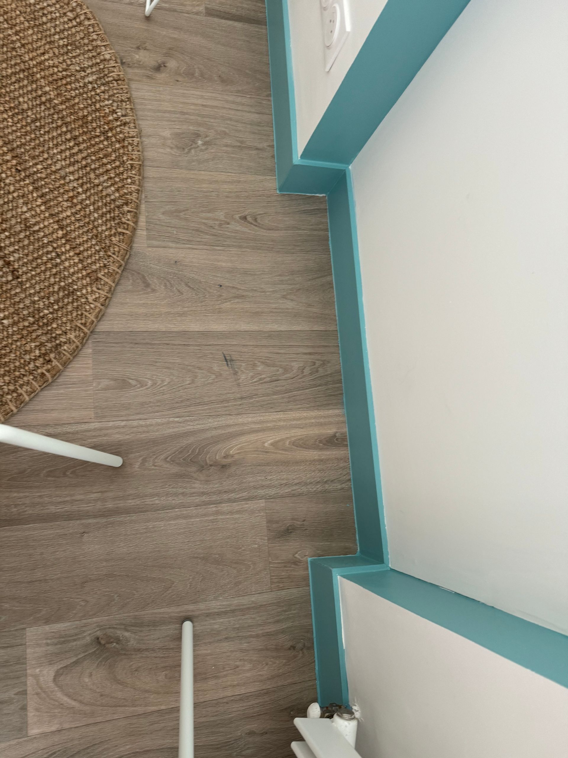 Vue en plongée d’un angle de petite pièce avec des boiseries, des murs, un parquet et une partie d’un tapis tissé et d’une chaise.