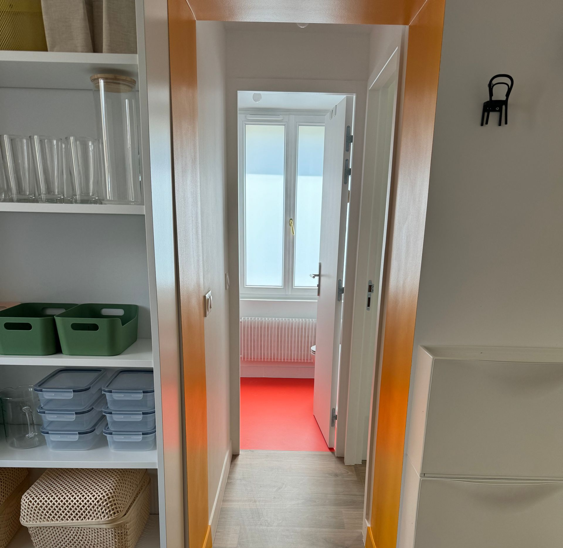 Couloir étroit avec des étagères à gauche, une porte ouverte au fond et un sol dans la pièce suivante.