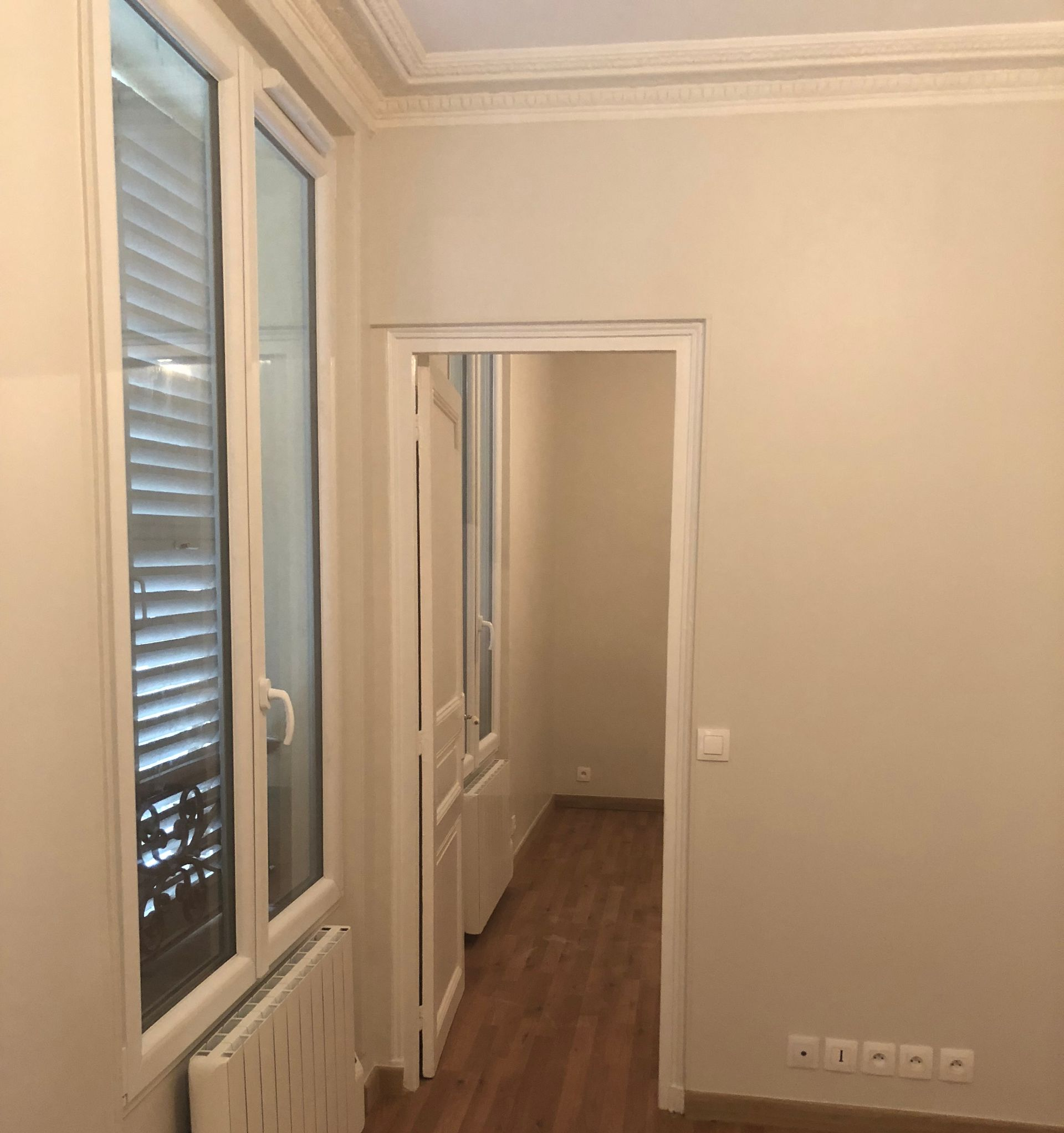 Couloir vide aux murs, au sol en bois, avec de hautes fenêtres et une porte donnant sur une autre pièce.
