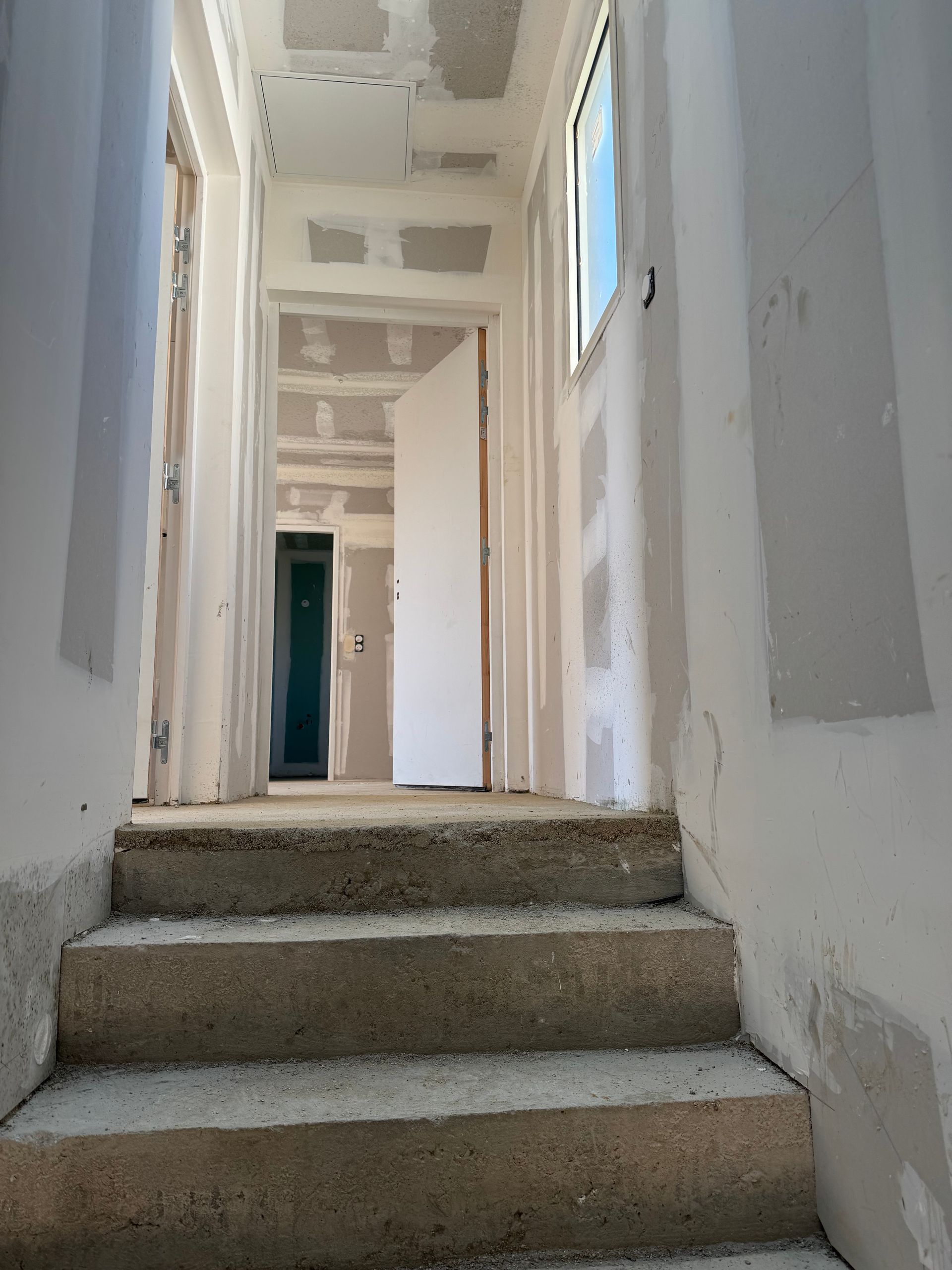 Couloir étroit, avec des plaques de plâtre apparentes et un petit escalier en béton menant à une porte.