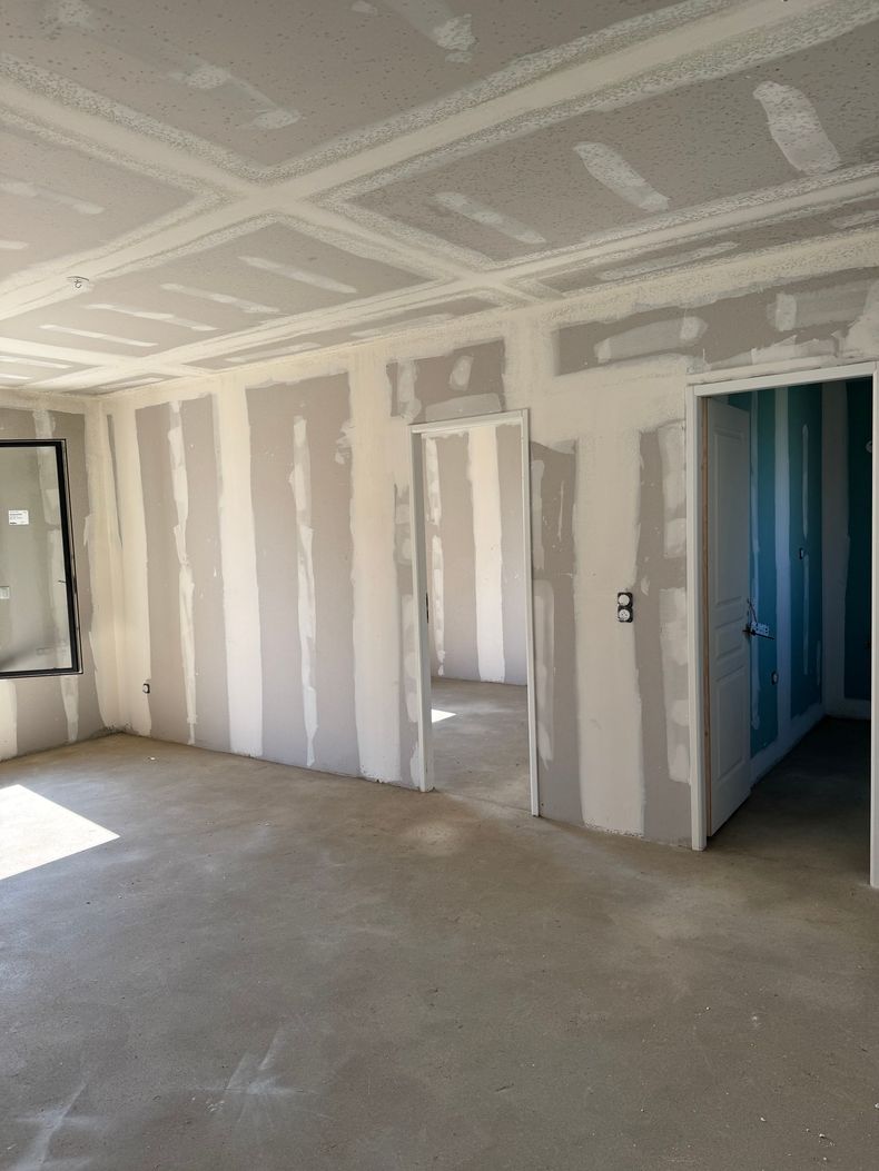 Pièce avec des joints de plaques de plâtre, un sous-plancher brut et des portes donnant sur un couloir et une pièce éclairée.