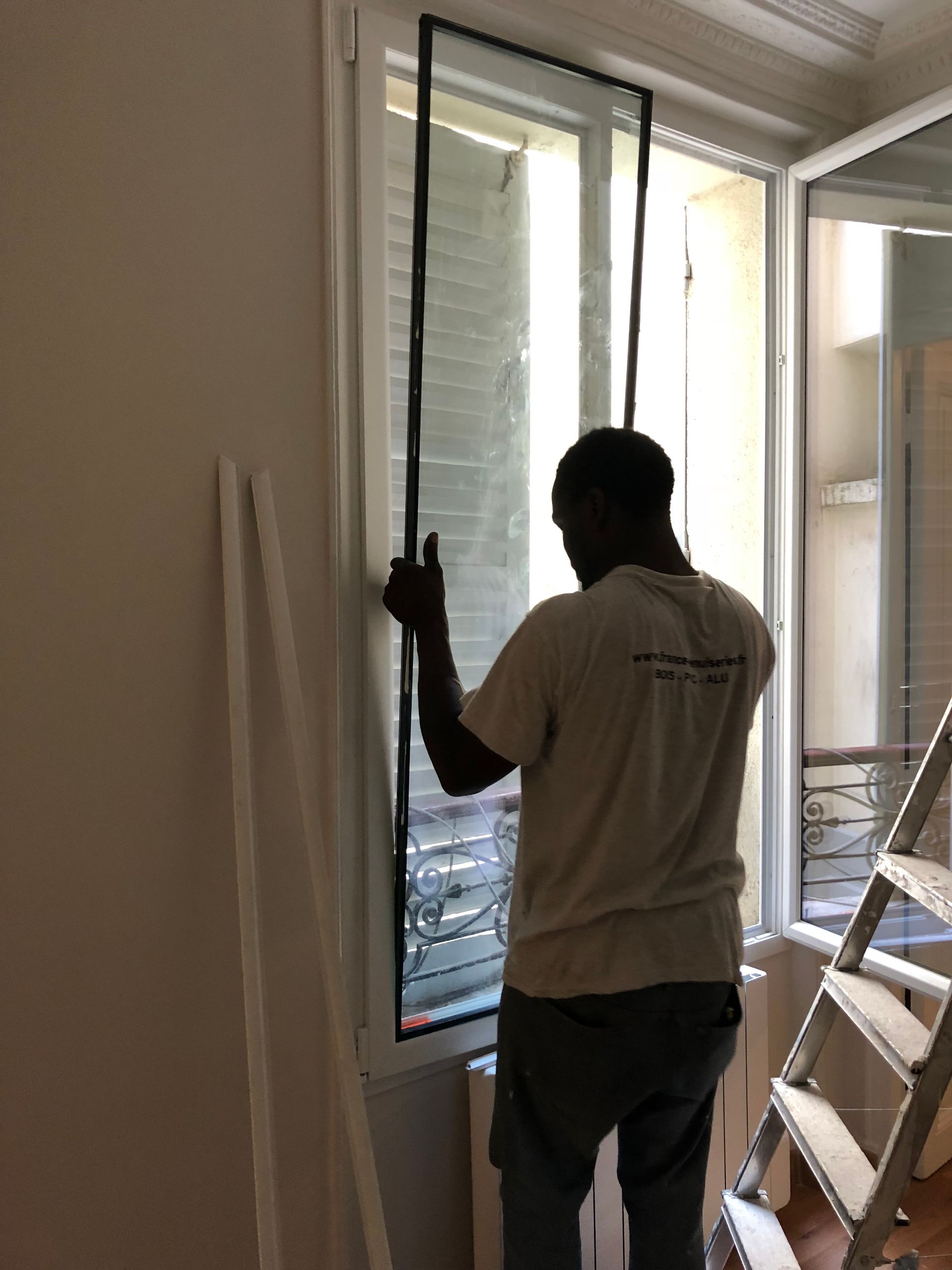 Une personne installe un store de fenêtre dans une pièce, à côté d’une échelle.