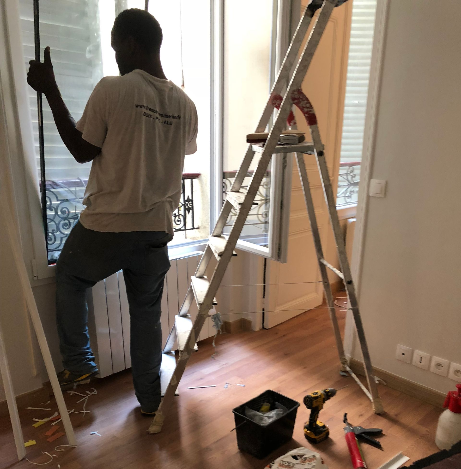 Personne sur une échelle travaillant près d’une fenêtre dans une pièce en rénovation, avec des outils et du matériel au sol.