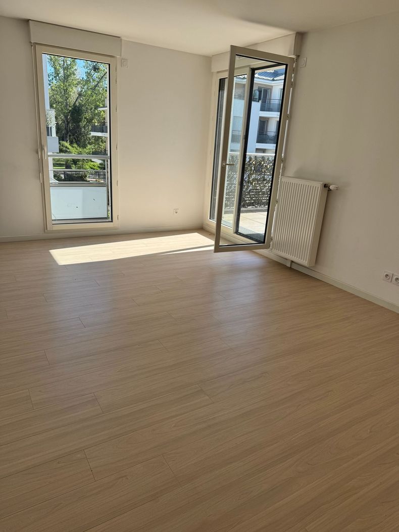 Pièce lumineuse et vide avec un parquet, des fenêtres et une porte ouverte donnant sur un balcon.