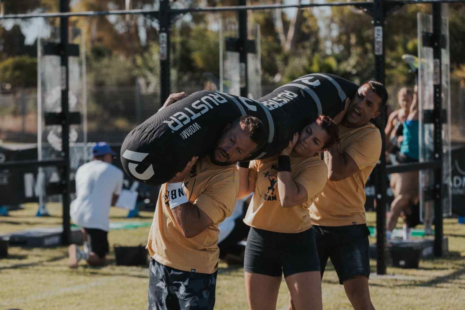 Dos personas cargando un gran obstáculo negro sobre sus hombros durante un evento de equipo al aire libre.