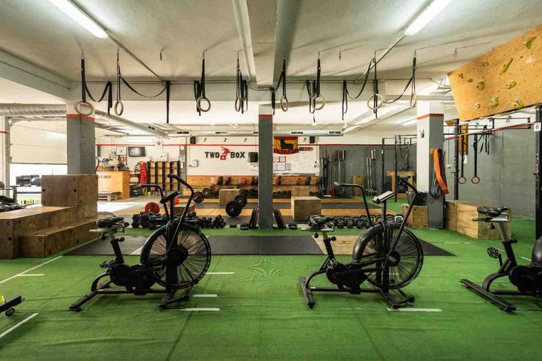 Gimnasio cubierto con máquinas de remo sobre césped artificial y correas de suspensión colgantes al fondo.