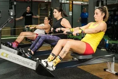 Hombre descansando en un banco de gimnasio junto a una barra y una bicicleta estática en un estudio de fitness.